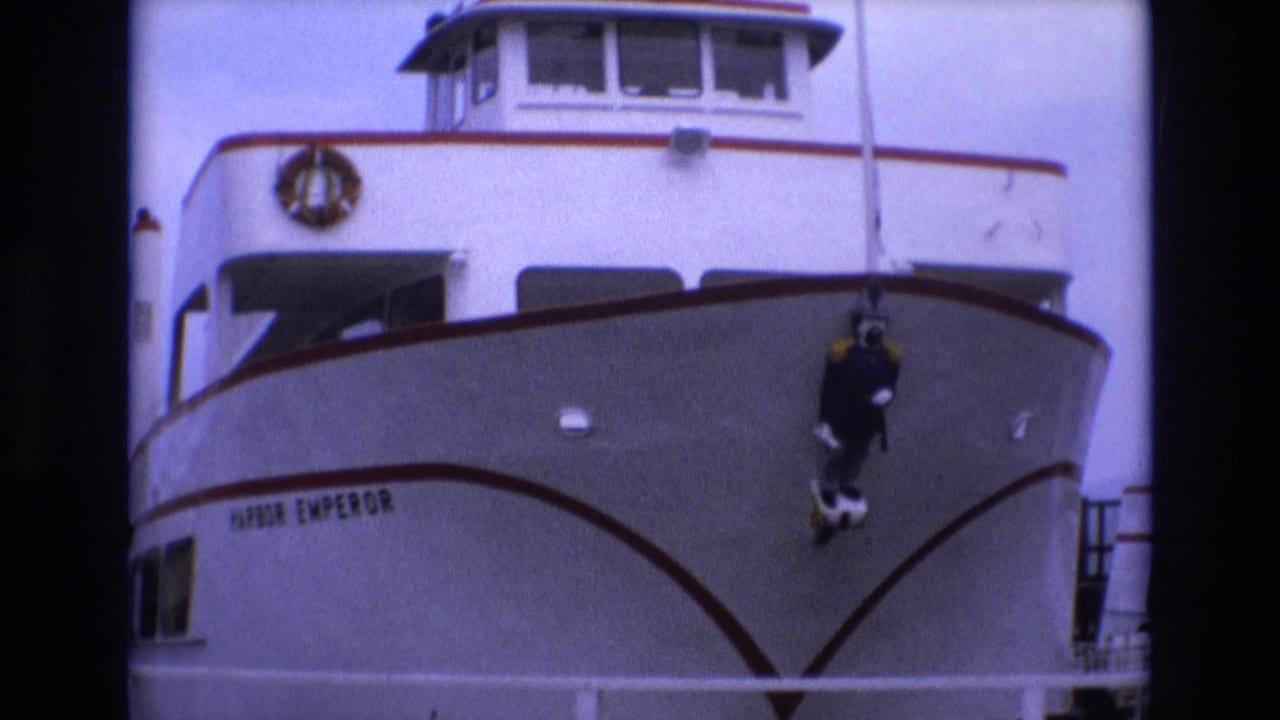 1973 San Francisco frame of the Harbor Emperor passenger vessel bow, with the vessel name visible and a dark-suited diver figure hanging near the center-right of the hull.