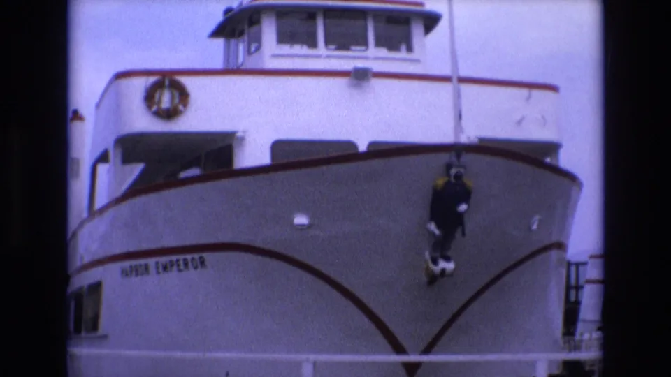 1973 San Francisco frame of the Harbor Emperor passenger vessel bow, with the vessel name visible and a dark-suited diver figure hanging near the center-right of the hull.