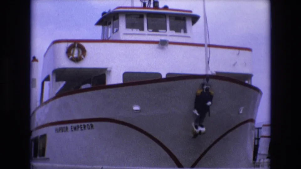 Final 1973 San Francisco frame showing the Harbor Emperor bow from below, with red trim, upper windows, life ring, and a dark diver figure hanging against the hull.