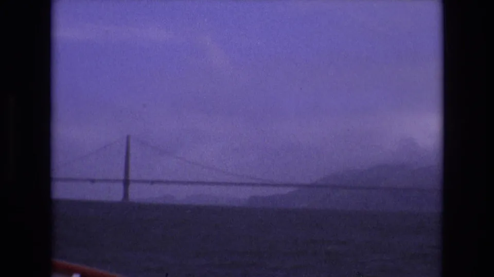 1973 San Francisco 8mm frame showing the Golden Gate Bridge stretching across the bay, with low hills in haze and a strip of boat railing at the bottom edge.