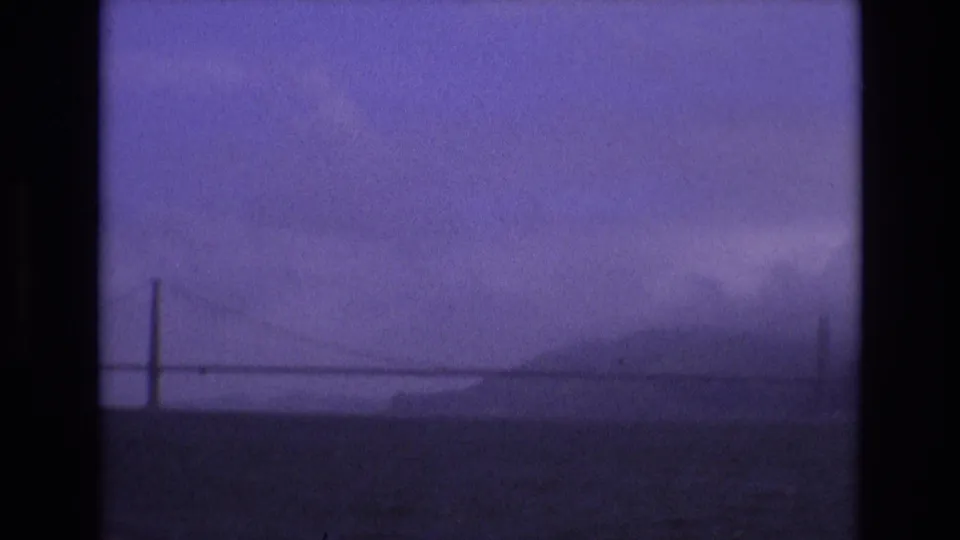 1973 San Francisco 8mm frame showing a faint Golden Gate Bridge silhouette, distant hills, fog, and dark bay water from a boat viewpoint.