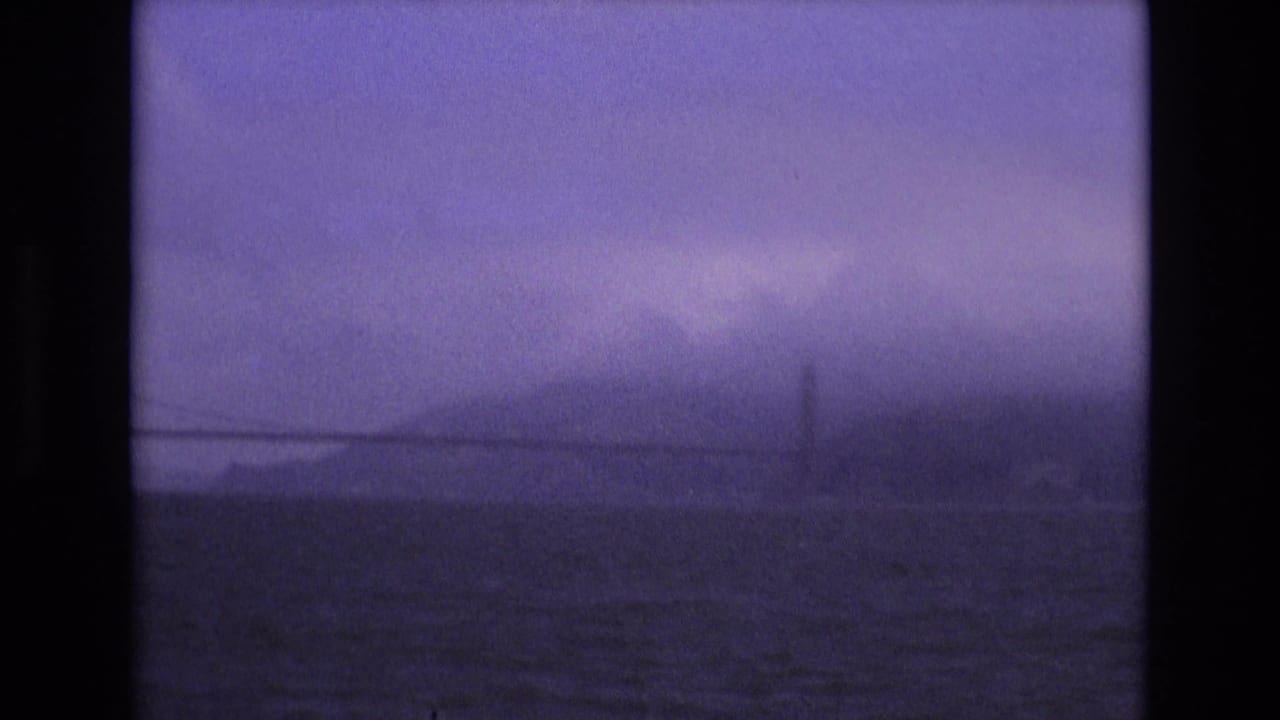 Faint bridge span and tower seen through purple-tinted fog above choppy water.