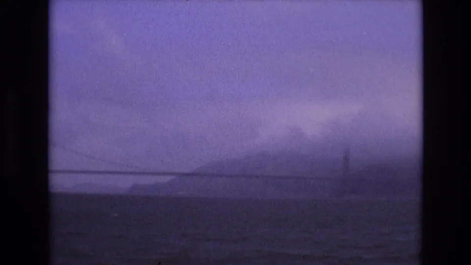1973 San Francisco 8mm frame showing a misted bridge line and hillside beyond choppy bay water, with heavy film grain and purple color shift.
