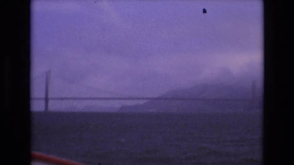 1973 San Francisco home-movie frame showing the Golden Gate Bridge tower and span emerging through fog, with hills and dark bay water below.
