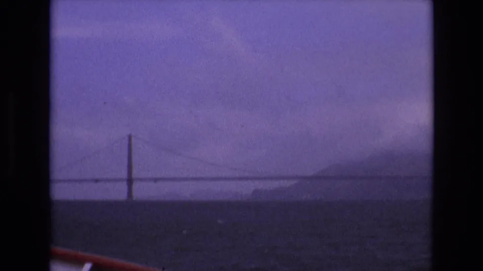 1973 8mm home-movie frame from San Francisco showing a distant long suspension bridge over bay water, tinted purple by aged film stock.