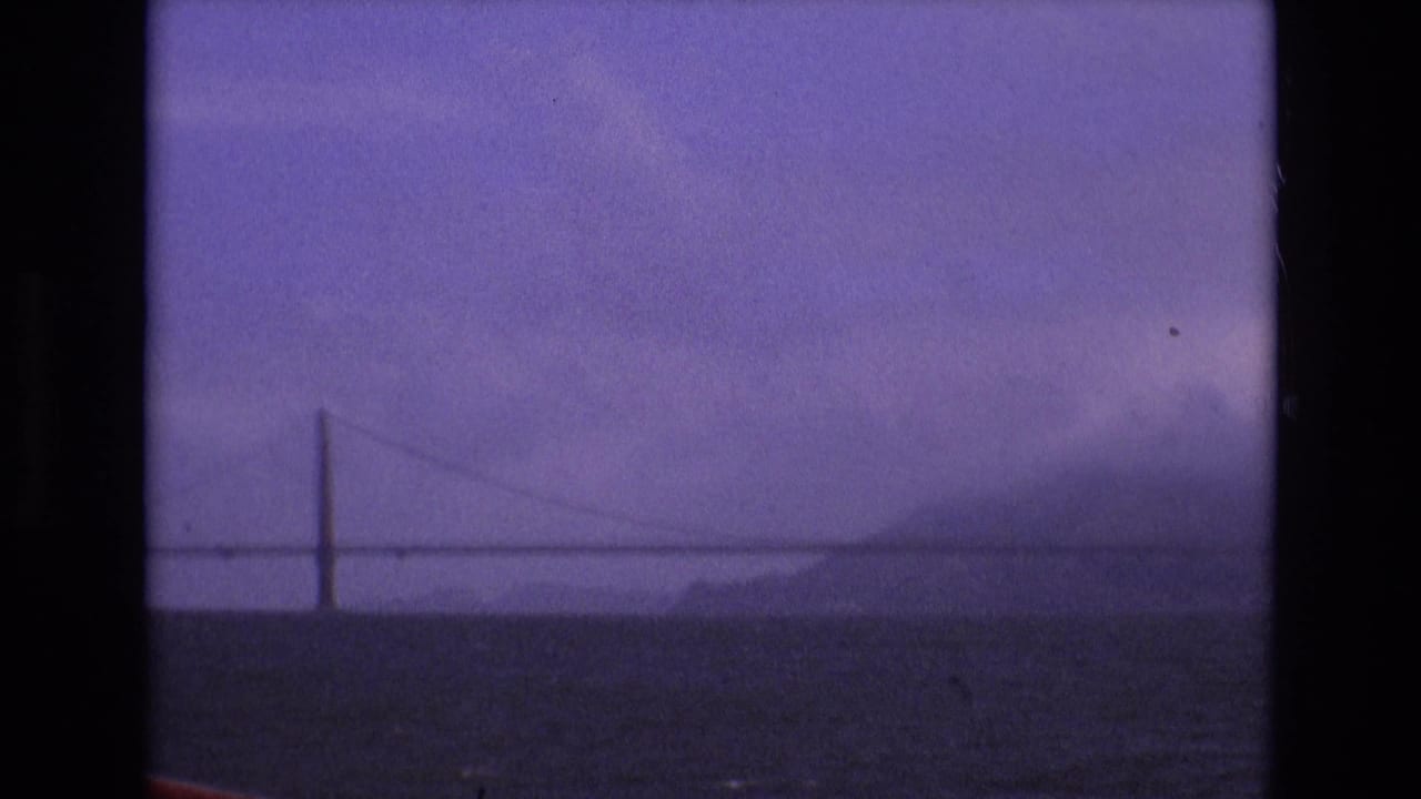 A distant San Francisco Bay suspension bridge appears through haze and soft focus, framed by 8mm film borders and purple color shift.