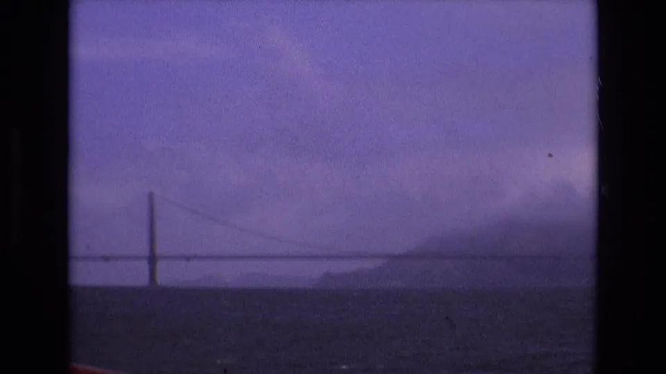 A distant San Francisco Bay suspension bridge appears through haze and soft focus, framed by 8mm film borders and purple color shift.