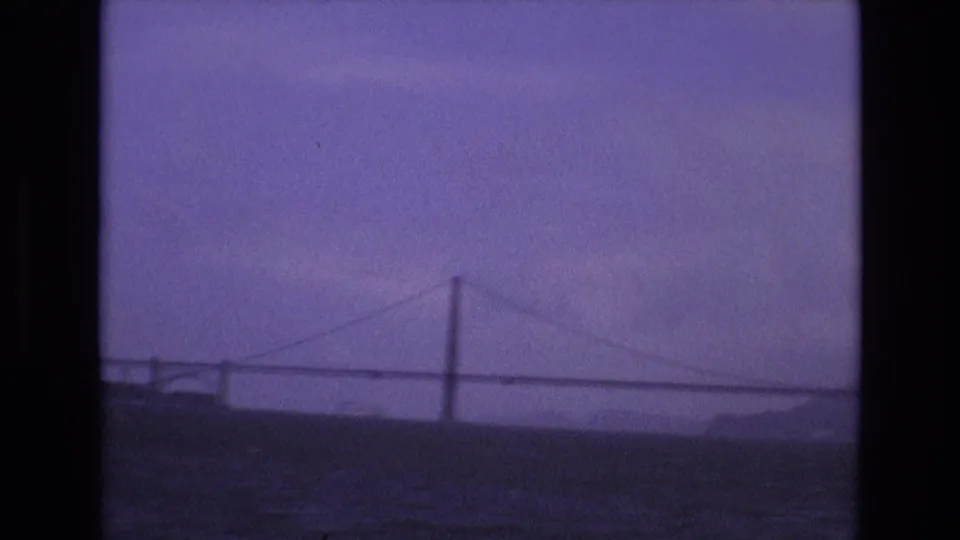 The moving camera catches the bridge at a slightly closer angle, with a shoreline edge at left and purple-tinted bay water below.