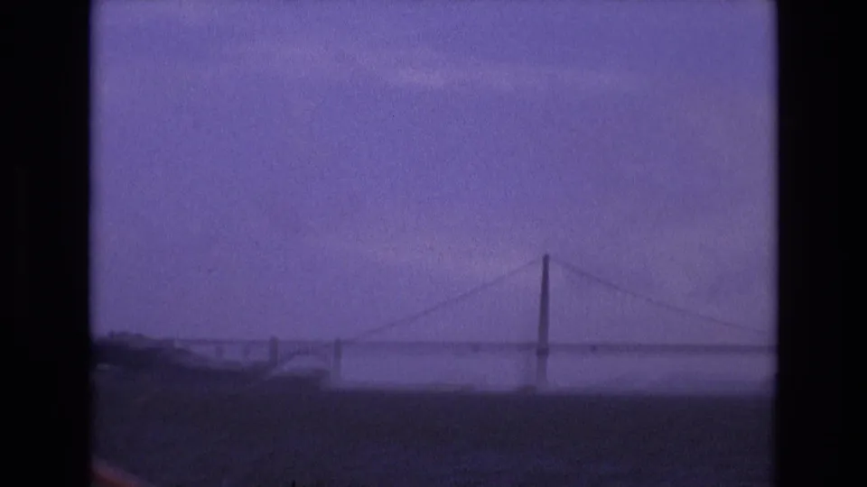 A bridge tower and suspension cables sit low on the horizon over San Francisco Bay water, with aged 8mm grain and a purple cast.