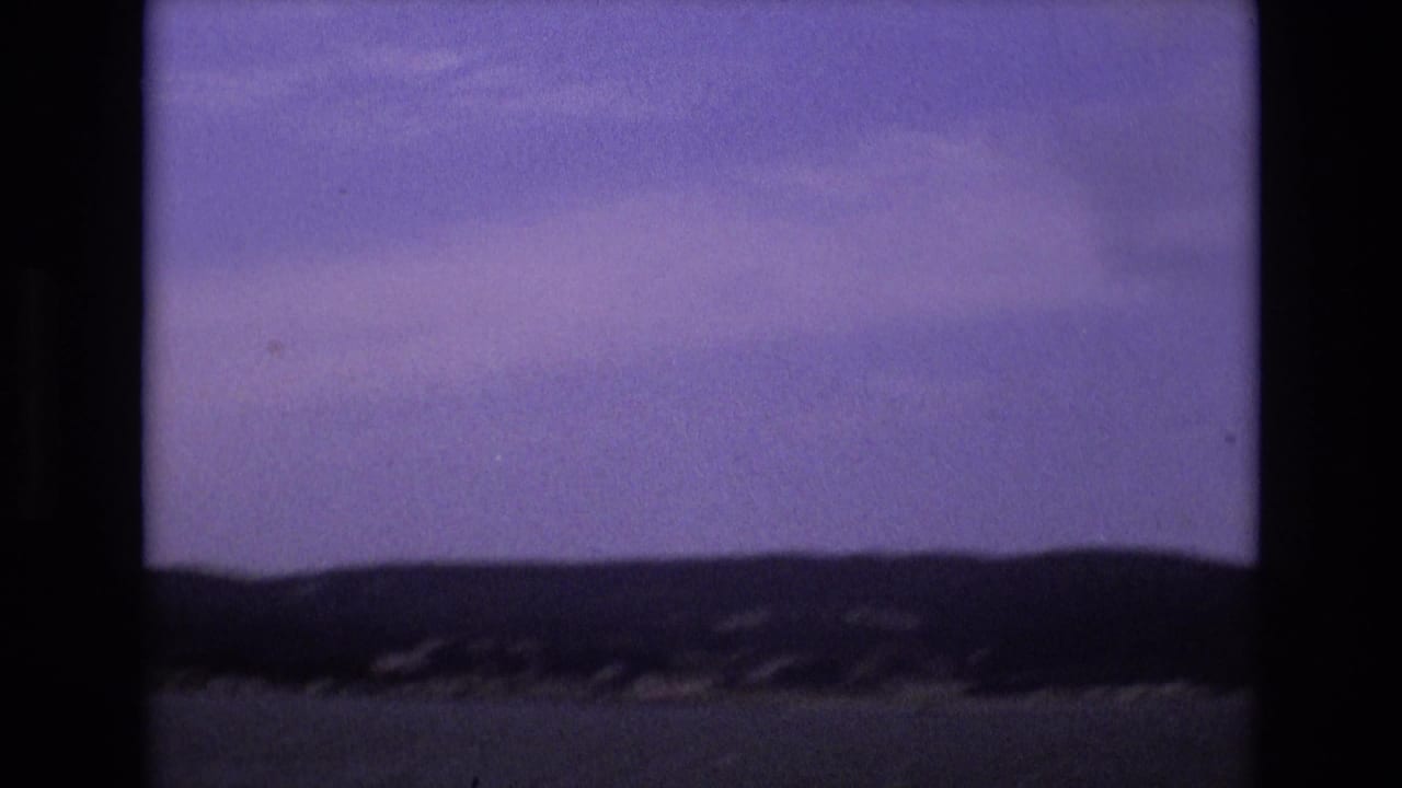 1973 San Francisco home-movie frame showing a dark distant shoreline and low hills across the bay beneath a muted, purple-tinted sky.