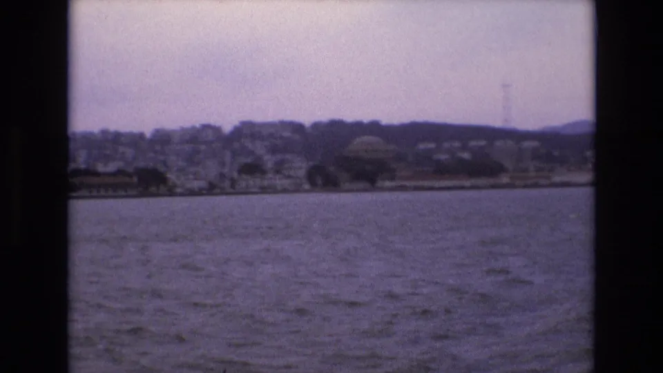 1973 San Francisco waterfront view from across the bay, with rippled water in the foreground and residential hills rising behind low shoreline structures.