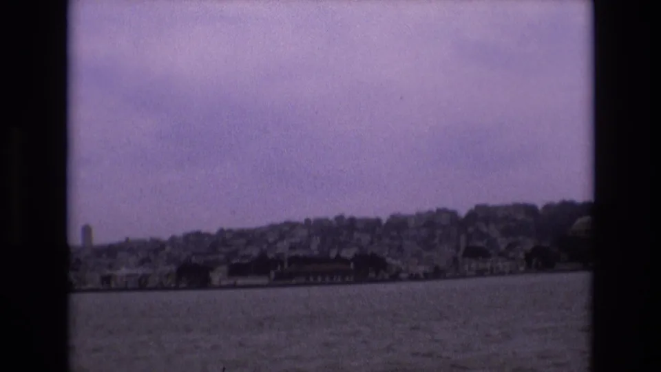 1973 San Francisco home-movie frame with open water in the foreground, a low shoreline, and densely built hillside neighborhoods under a pale sky.