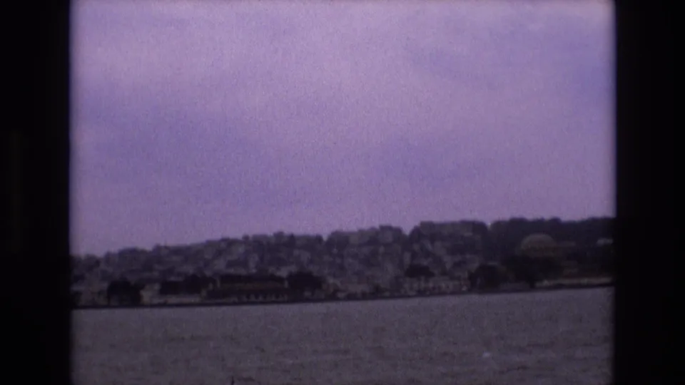 1973 8mm view from a boat looking across bay water toward San Francisco hillside buildings under a hazy purple-tinted sky.