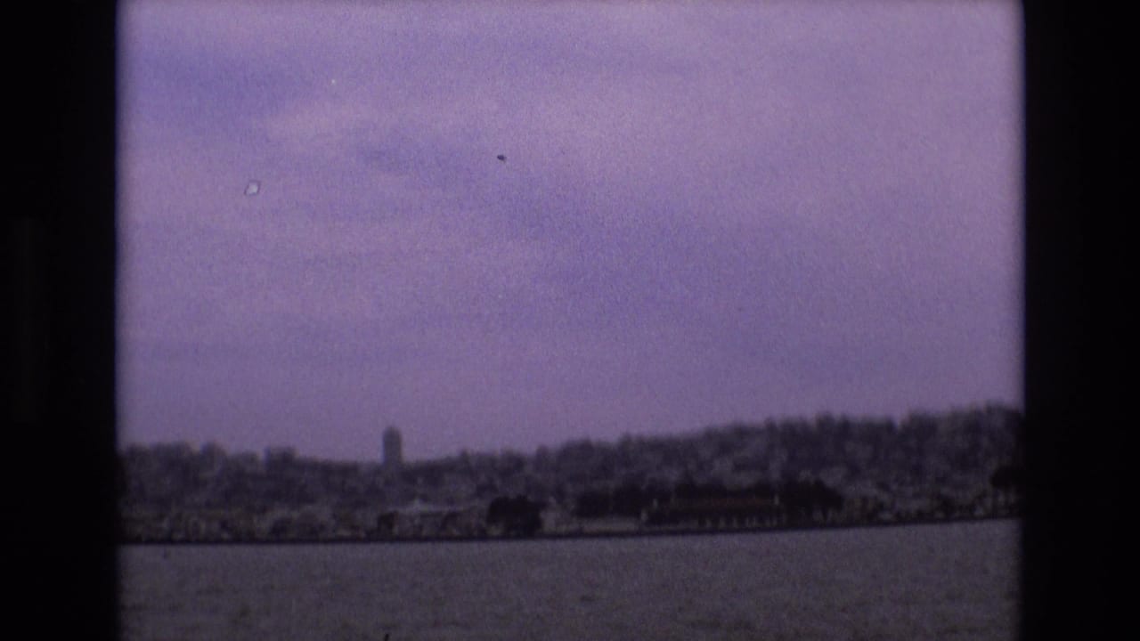 1973 8mm home-movie frame showing San Francisco shoreline and hills seen across water, with soft focus, film grain, and dark frame edges.