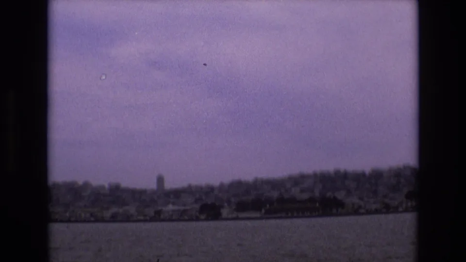 1973 8mm home-movie frame showing San Francisco shoreline and hills seen across water, with soft focus, film grain, and dark frame edges.