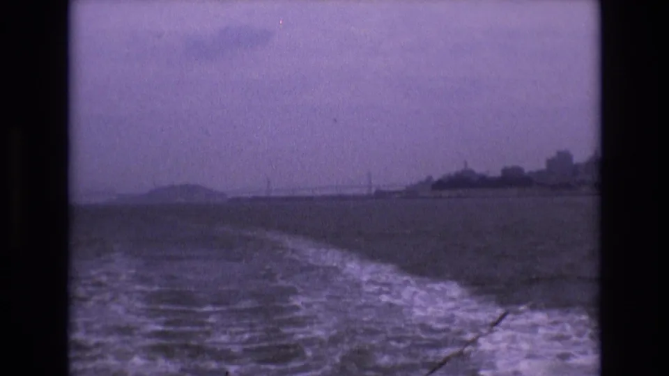 From a moving boat on San Francisco Bay, the frame looks back across a foamy wake toward a hazy shoreline, low hills or bridge forms, and distant urban shapes.
