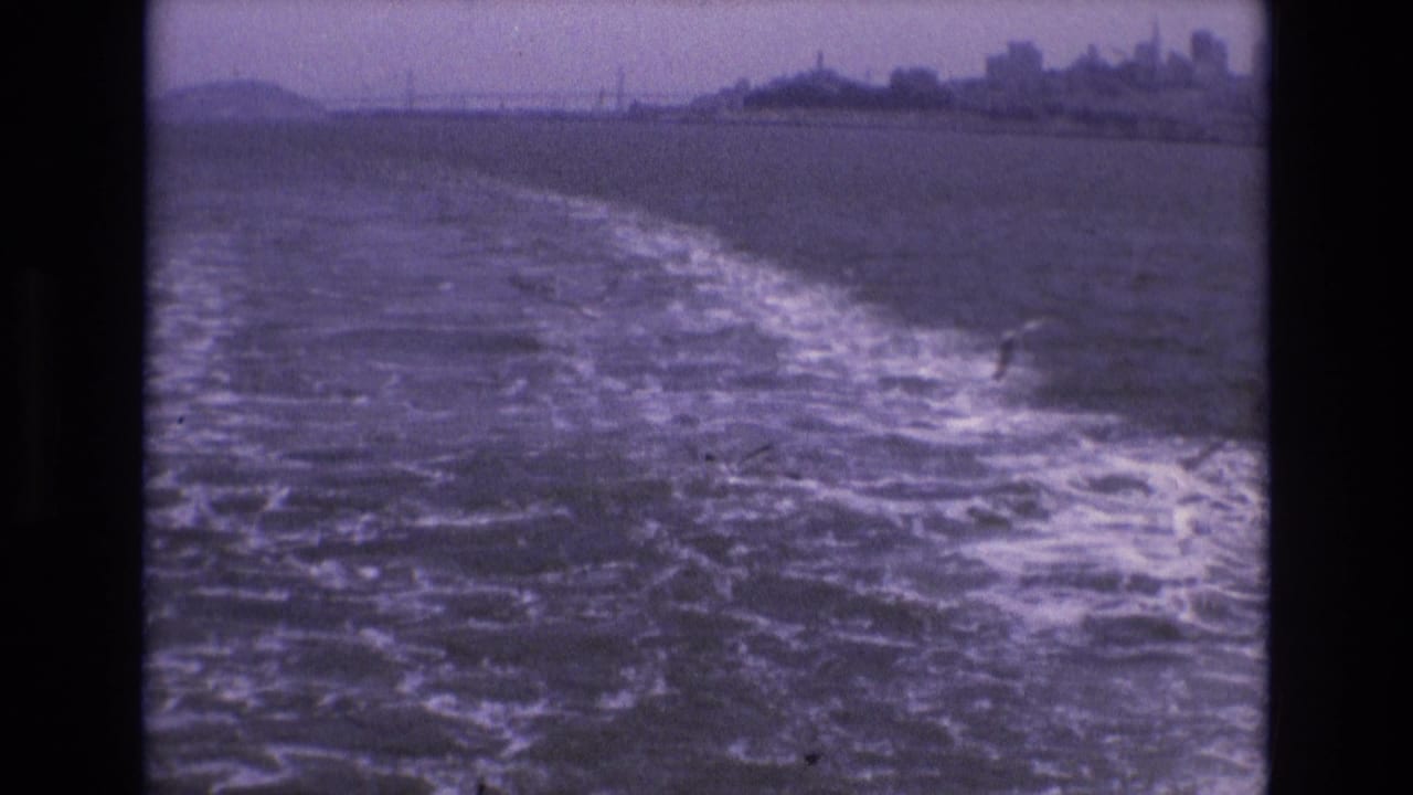 1973 8mm home-movie view from a moving boat on San Francisco Bay, with foamy wake, choppy water, distant city skyline, and bridge on the horizon.