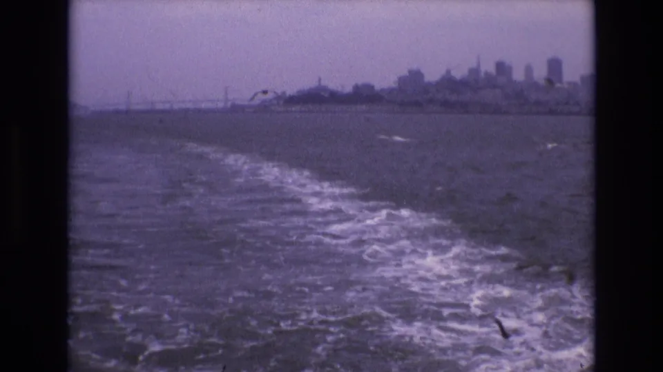 Soft, purple-tinted 8mm view of San Francisco Bay from a moving boat, with wake, gulls, distant skyline, and bridge under an overcast sky.