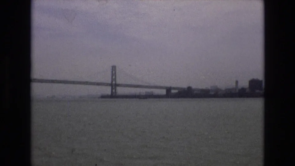 1973 home-movie frame of the Golden Gate Bridge from a boat on San Francisco Bay, with low shoreline buildings and muted overcast light.