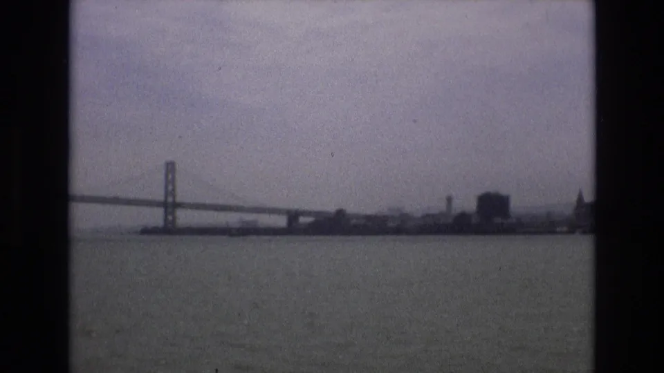 1973 8mm home-movie frame showing the Bay Bridge and a distant San Francisco waterfront skyline across gray bay water under a hazy sky.
