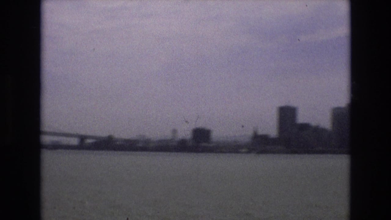 1973 San Francisco waterfront view with part of the Bay Bridge, bay water, and distant high-rises softened by haze and 8mm film grain.