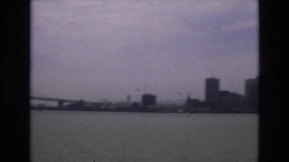 1973 San Francisco waterfront view with part of the Bay Bridge, bay water, and distant high-rises softened by haze and 8mm film grain.