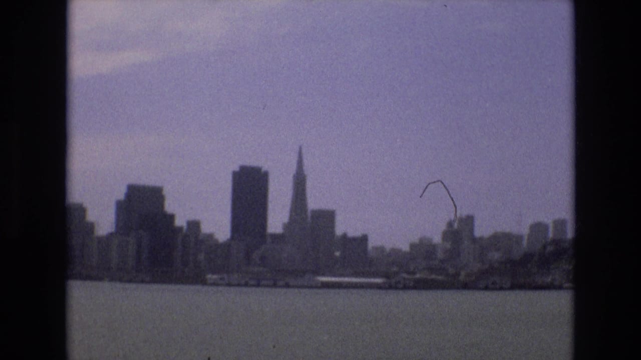 The San Francisco skyline appears beyond calm bay water in a 1973 8mm frame, with the Transamerica Pyramid and clustered downtown towers under a muted sky.