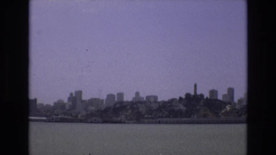 A wide 1973 8mm home-movie frame shows San Francisco waterfront and skyline forms across the bay, with open water in the foreground and a pale hazy sky above.
