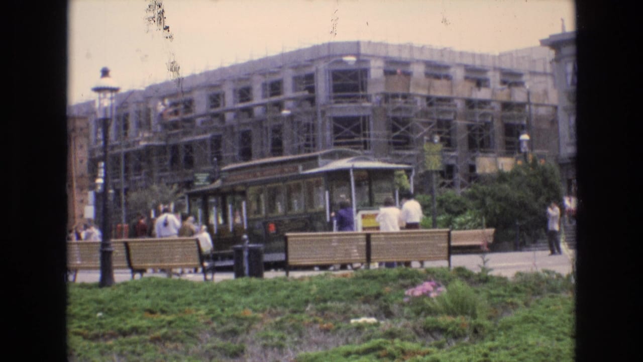 Streetcar stopped near benches, lampposts, pedestrians, greenery, and a scaffolded building.