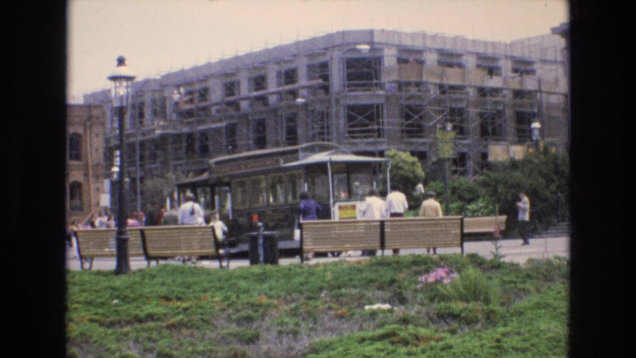 A San Francisco streetcar pauses at a busy stop in 1973, with people nearby, benches in front, and a scaffolded building filling the background.