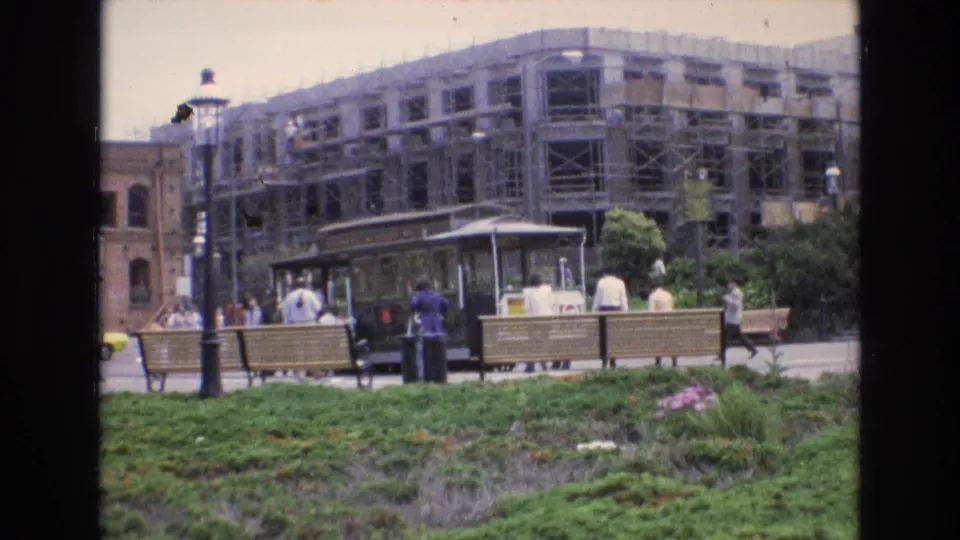 The 8mm frame shows a 1973 San Francisco streetcar stop with pedestrians, benches, street lamps, planted foreground, and visible construction scaffolding behind.