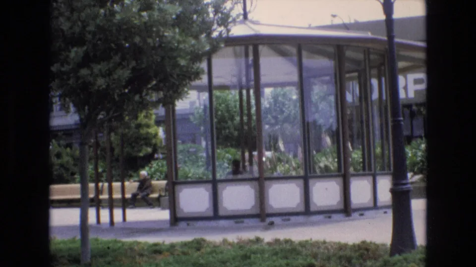 Later 1973 San Francisco frame shifts toward a glass-enclosed pavilion, with a seated pedestrian, benches, trees, planting, and street lamps nearby.