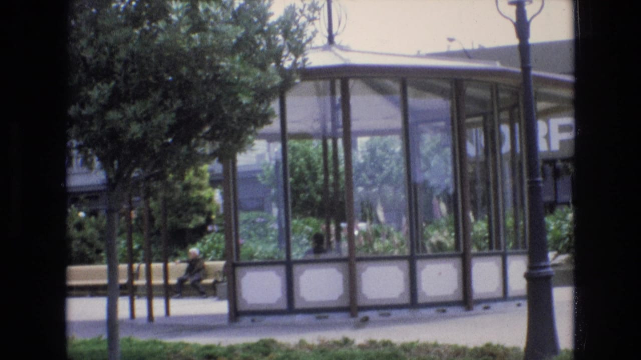 Glass pavilion in a park with trees, a bench, and a seated person nearby.