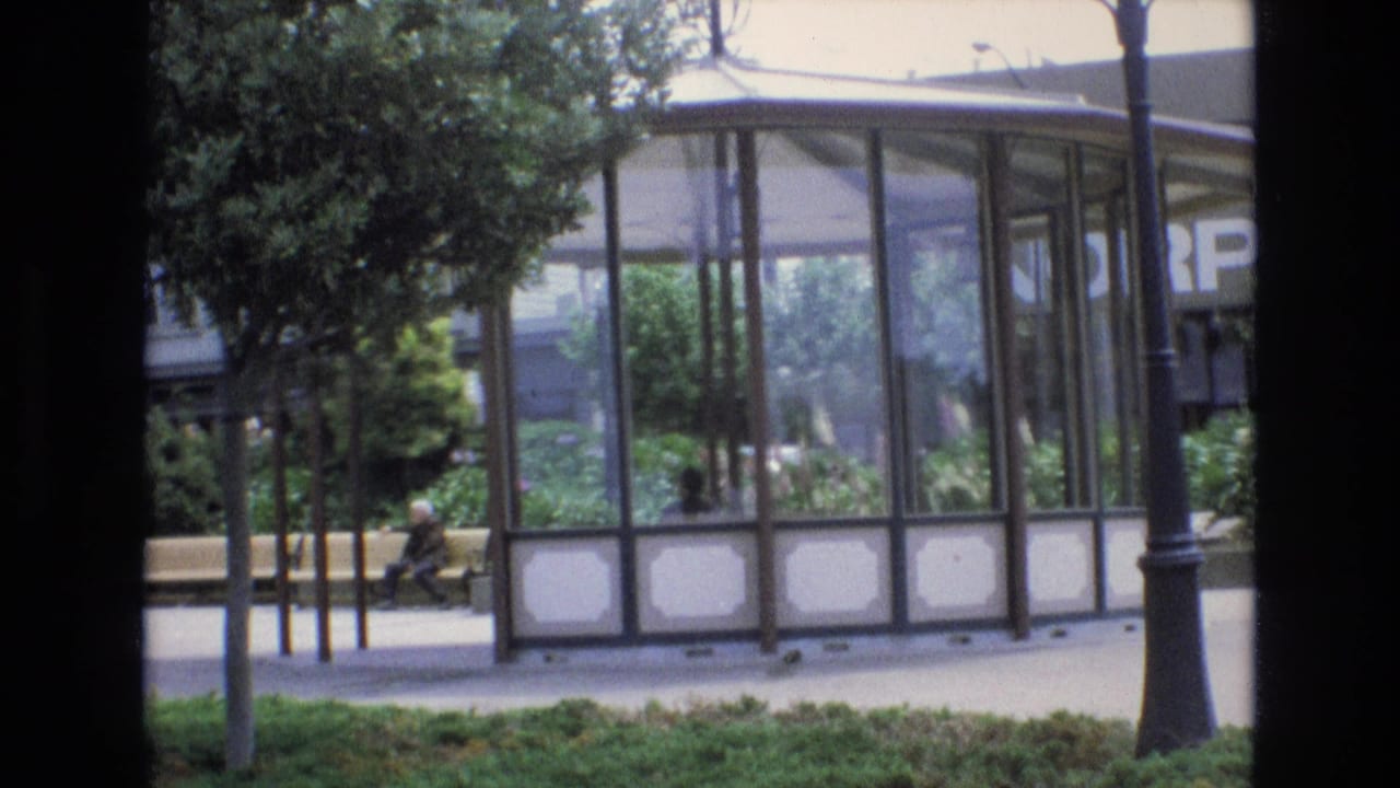 1973 San Francisco home-movie frame with a glass shelter or pavilion framed by trees, a lamp post, planting beds, and a person sitting on a bench.