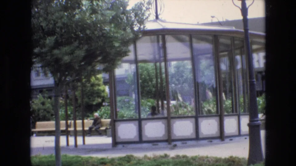 Slightly tilted 1973 San Francisco 8mm frame showing a glass park pavilion, leafy trees, a bench with a seated figure, shrubs, and a paved walkway.