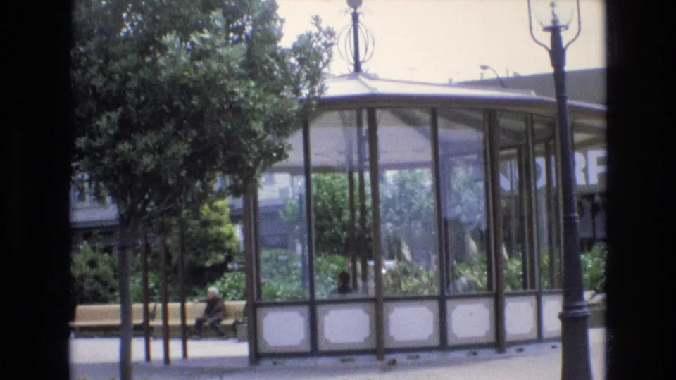 1973 San Francisco park frame showing a glass-enclosed pavilion, dense greenery, lamp post, walkway, and a person seated on a bench near the trees.