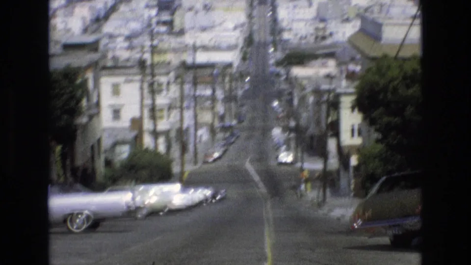Soft 1973 San Francisco 8mm frame with parked classic cars at the base of a steep residential street lined by buildings and trees.