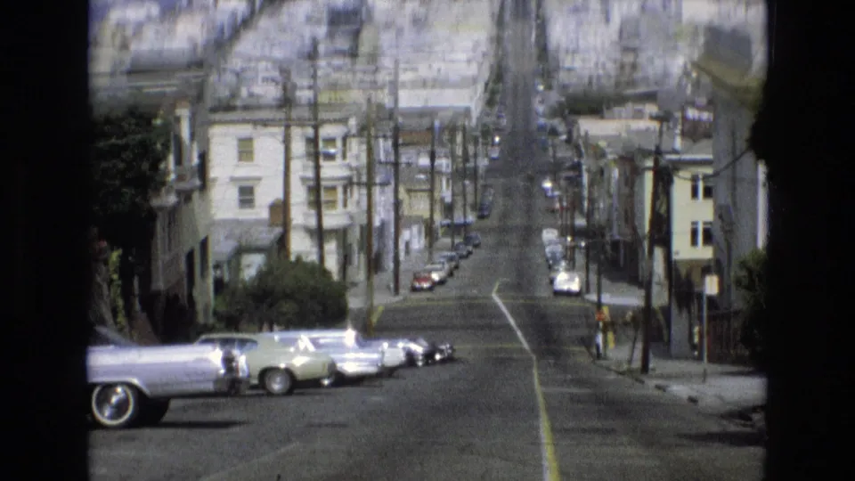 1973 San Francisco home-movie frame looking up a steep street with classic cars, dense residential buildings, overhead wires, and a distant person near the curb.