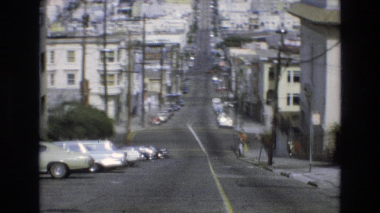 Hilly residential street with parked cars, buildings, and a small pedestrian on the sidewalk.