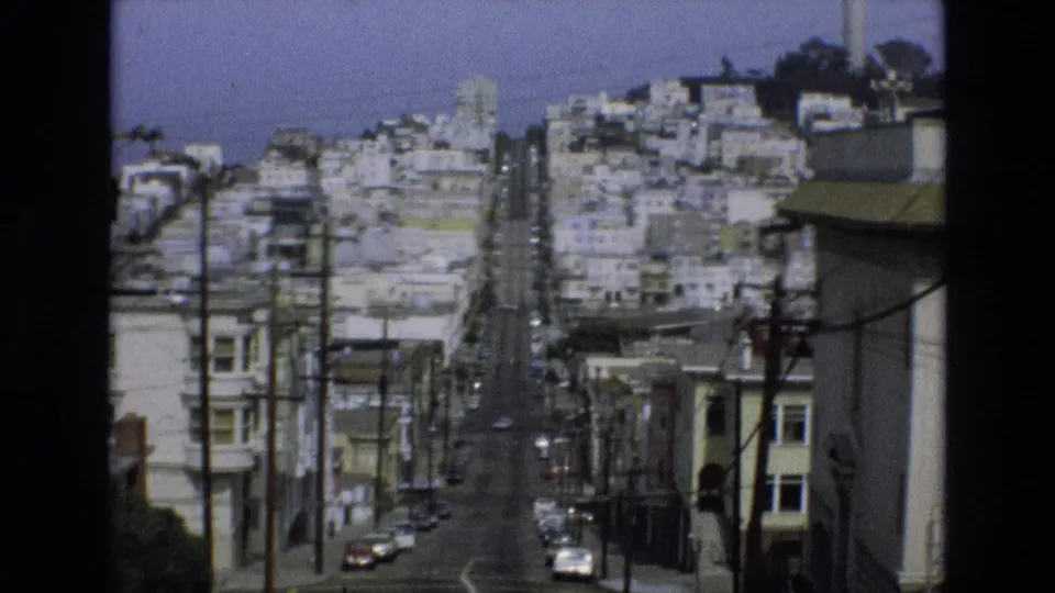1973 San Francisco home-movie frame with a long downhill view through dense residential buildings, utility poles, wires, and cars on the sloped street.