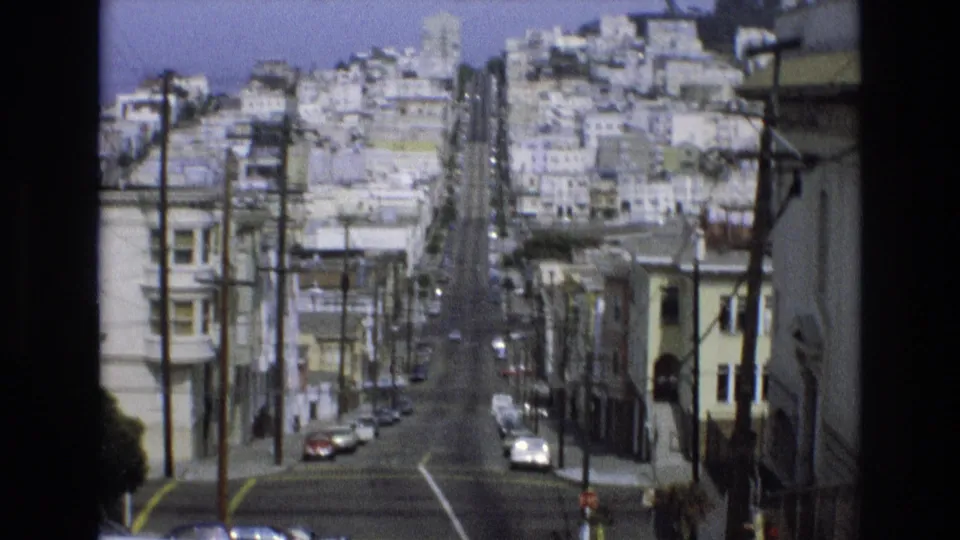 1973 San Francisco residential hill street seen from above, with classic cars, pale buildings, utility poles, overhead wires, and a small sidewalk figure.