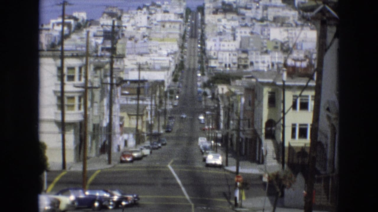 1973 San Francisco home-movie frame showing a steep downhill residential street with older parked cars, utility poles, hillside buildings, and small pedestrians near the sidewalks.