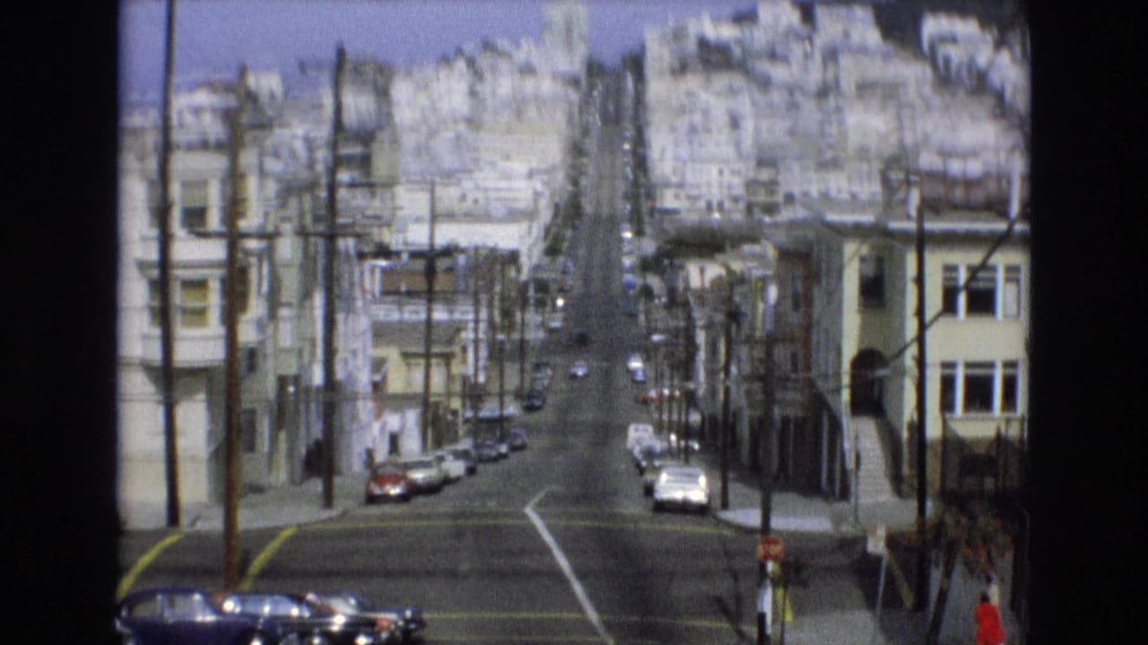 Downhill city street with parked cars, overhead wires, dense buildings, and pedestrians near the sidewalk.
