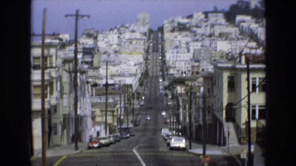 Soft 8mm frame of a steep San Francisco residential street in 1973, with hillside housing, utility poles, parked cars, and a broad downhill view through the neighborhood.