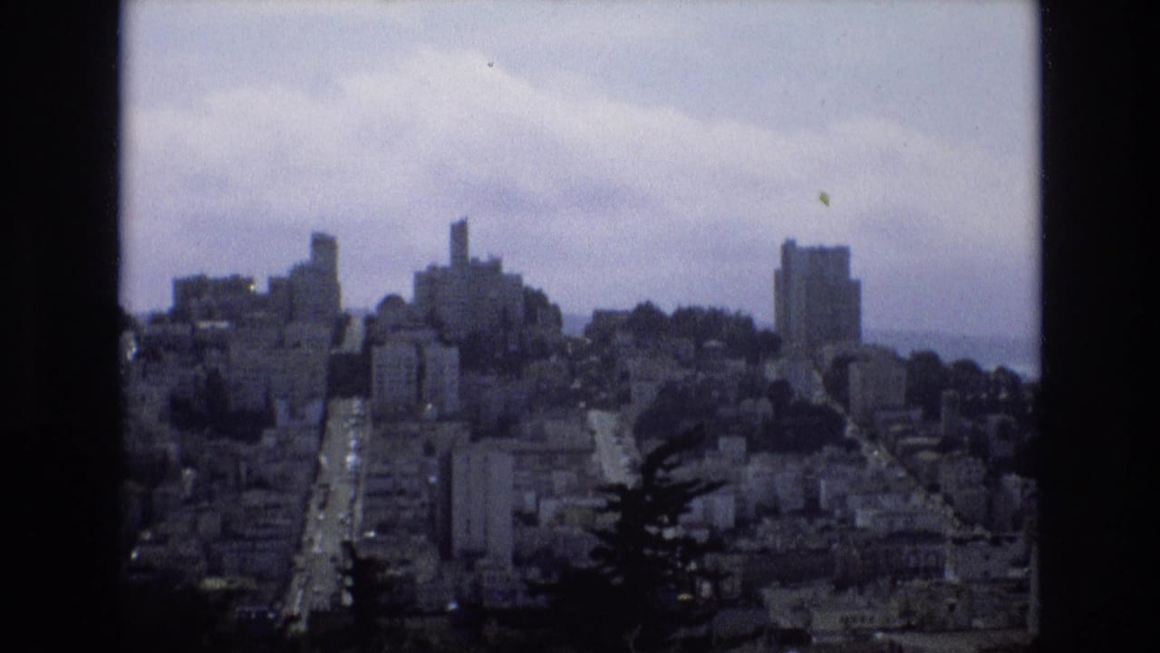 1973 8mm view of San Francisco from a high vantage point, with dense hillside buildings, a central road, dark foreground trees, and a cloudy sky.