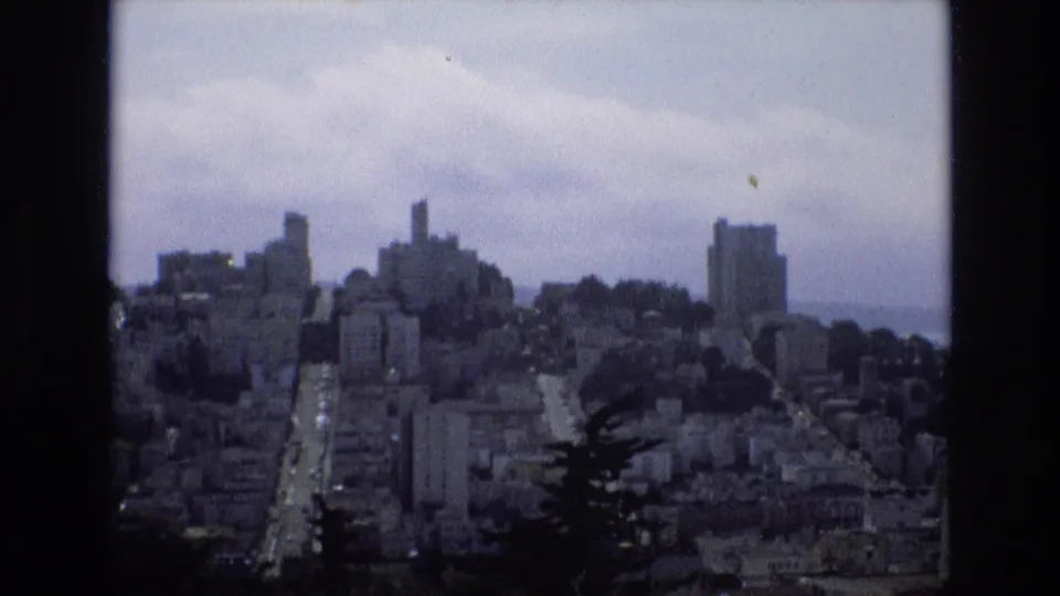 1973 8mm view of San Francisco from a high vantage point, with dense hillside buildings, a central road, dark foreground trees, and a cloudy sky.