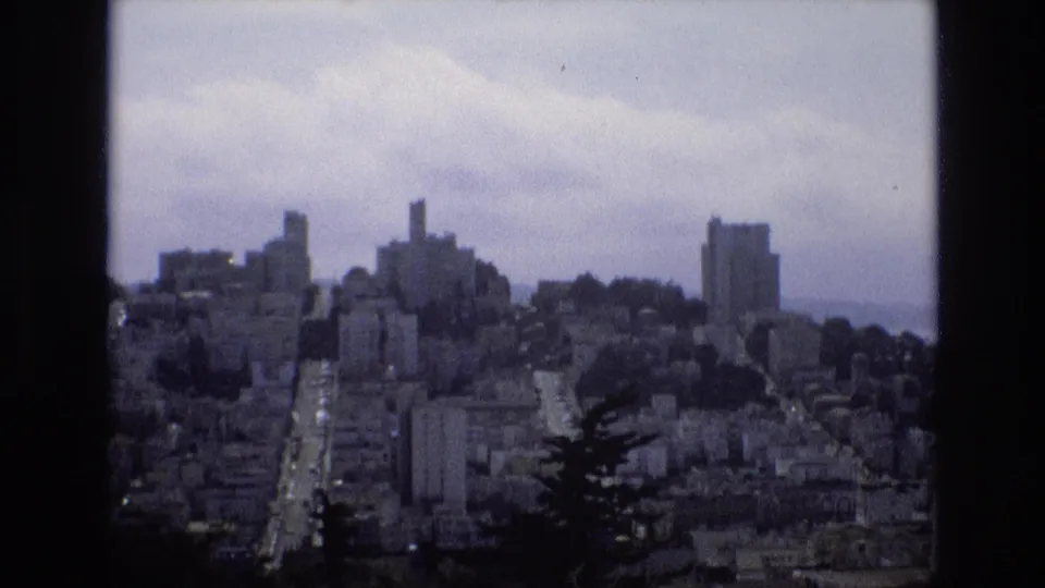 San Francisco appears in a soft 1973 8mm frame, showing mid-rise and taller buildings across hills with a bright roadway running toward the distance.