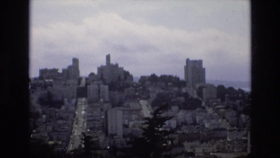 A wide 1973 home-movie frame looks across San Francisco's hills, showing dense buildings, dark foreground trees, a visible road corridor, and pale clouds.
