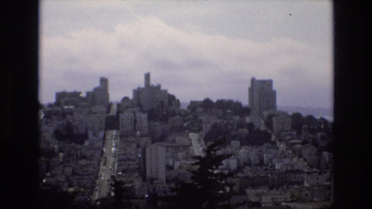 1973 8mm home-movie frame of San Francisco hills and dense buildings under a cloudy sky, seen from an elevated vantage point with dark trees in the foreground.