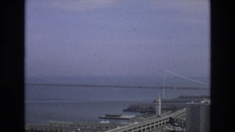 1973 8mm frame of a San Francisco bay bridge view, with water, a long span, waterfront structures, and roads seen from a high overlook.